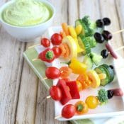 5 vegetable kabobs laying across a rectangular white platter on green and white cloth with veggie dip in background.