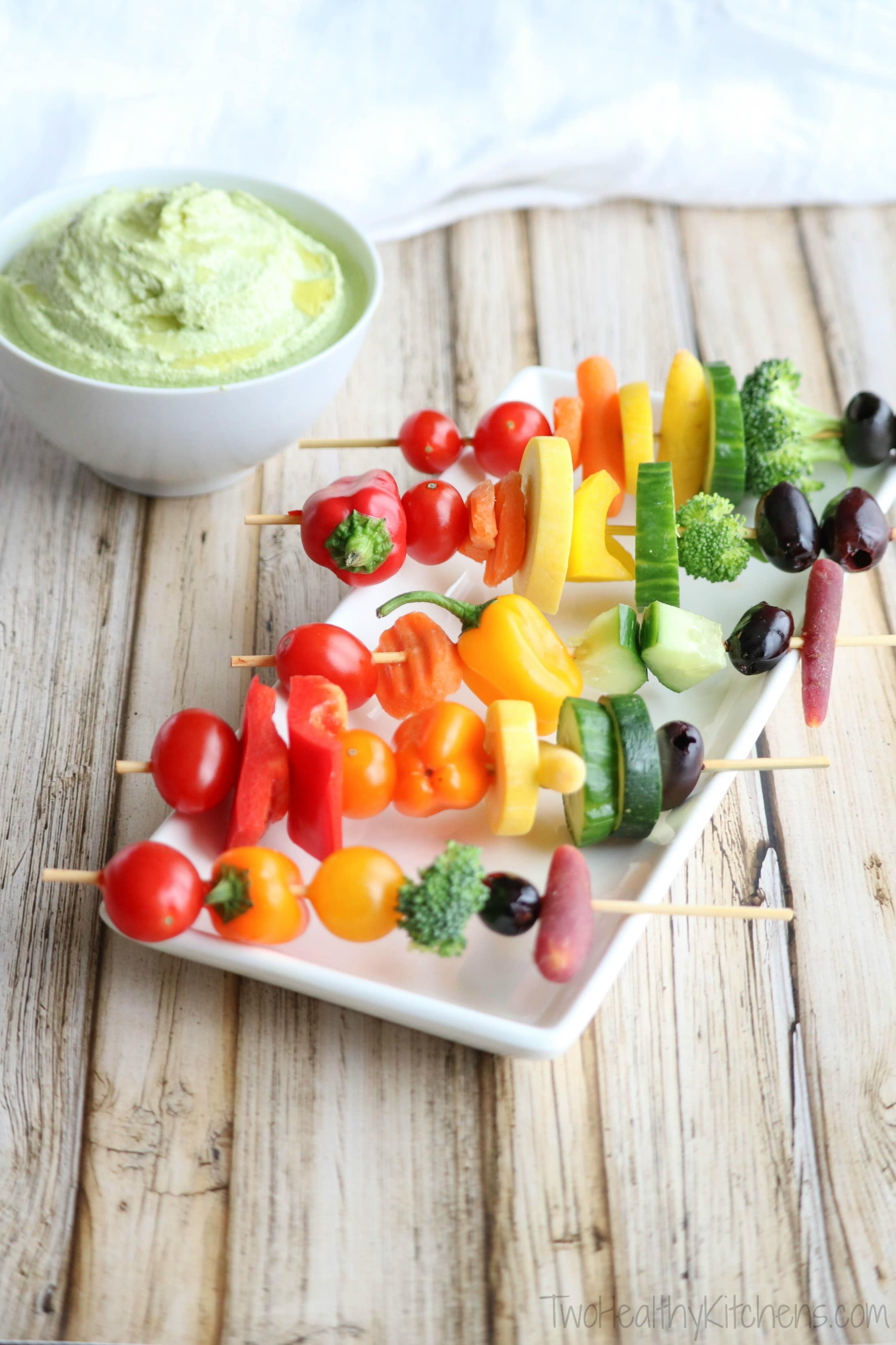 Skewered veggies in rainbow-colored rows on serving plate with green vegetable dip in bowl in background.