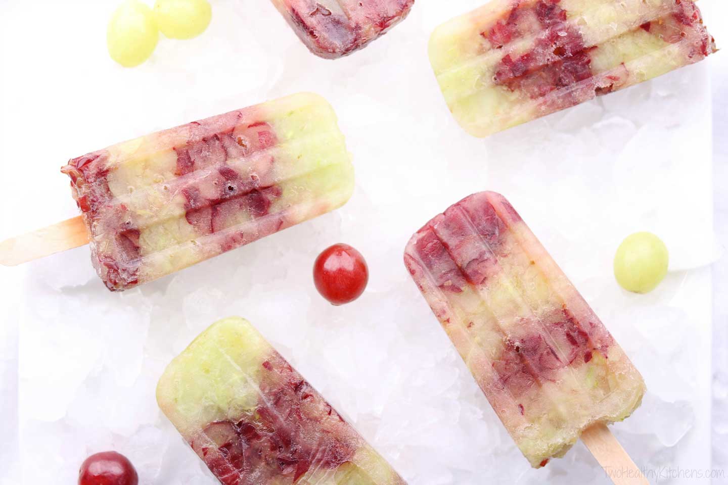 Several striped, frozen grape pops on ice with single green and red grapes scattered amongst.