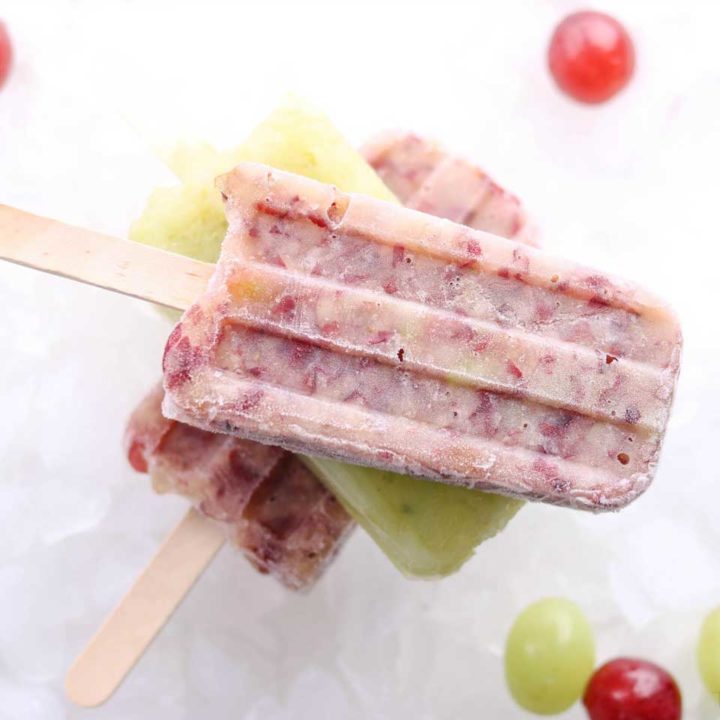 Flatlay of stacked pops laying on top of each other, a red one on top, with green and red grapes scattered nearby.