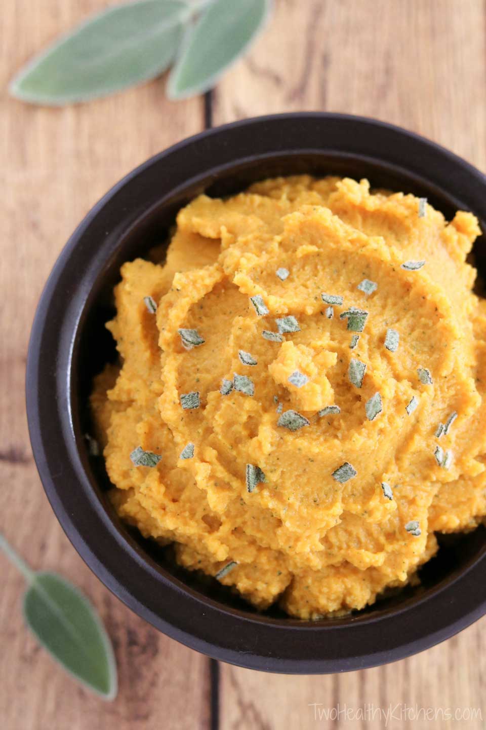 Overhead photo of Pumpkin Hummus in a little brown bowl, sprinkled with bits of fresh sage and with whole sage leaves nearby.