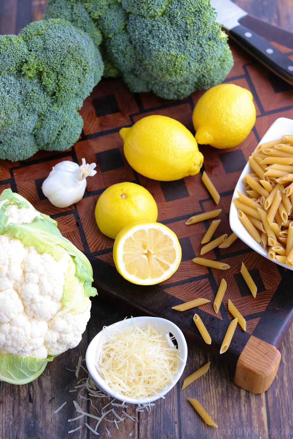 overhead of the ingredients for this recipe, including a head of cauliflower and two bunches of broccoli, several lemons, uncooked penne, a little bowl of parmesan shreds, and a head of garlic
