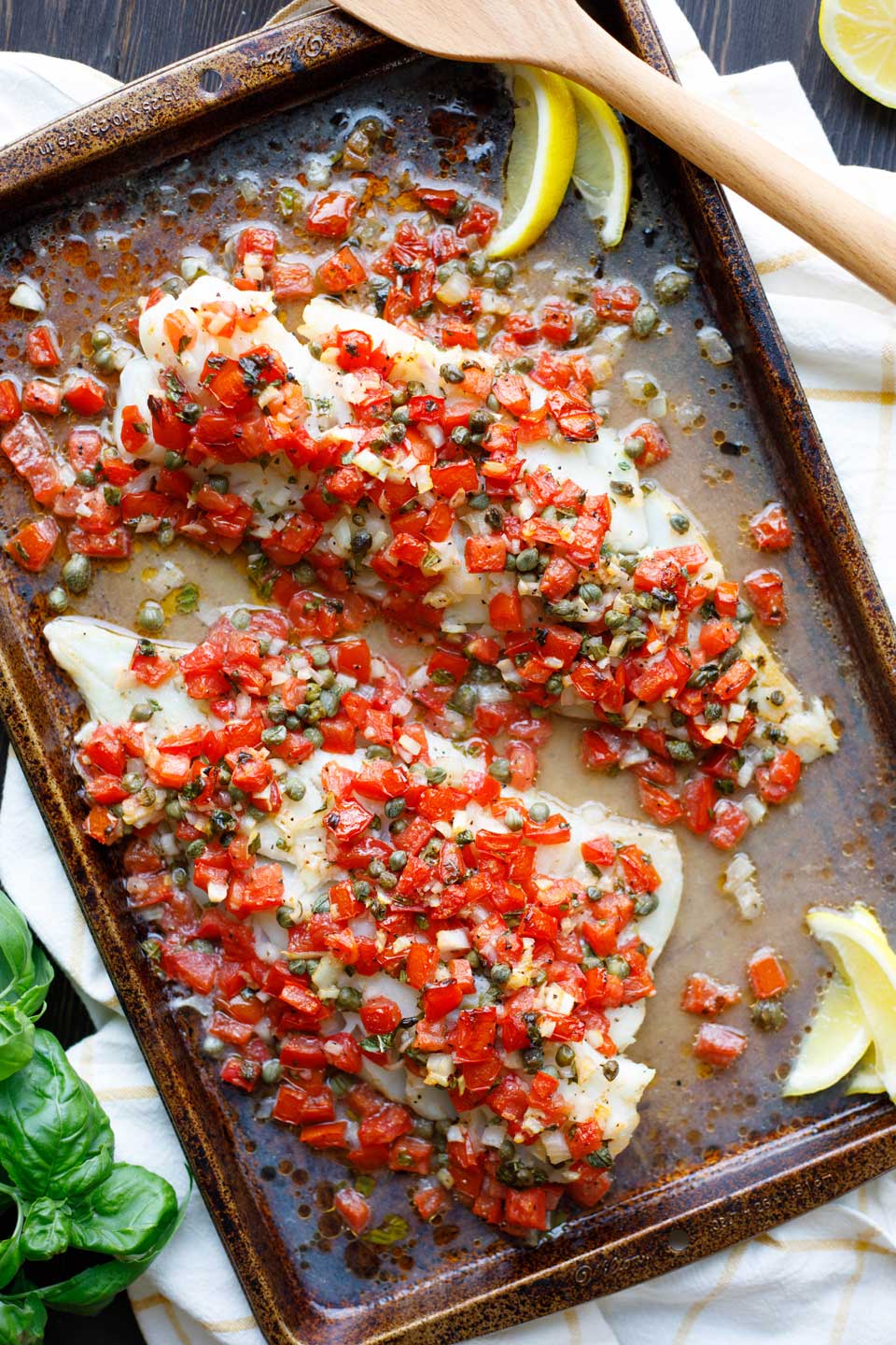 Overhead photo of two broiled fish fillets on a rimmed baking sheet, topped with the recipe's tomato, caper, and basil topping mixture, and with lemon wedges and fresh basil around the edges. 