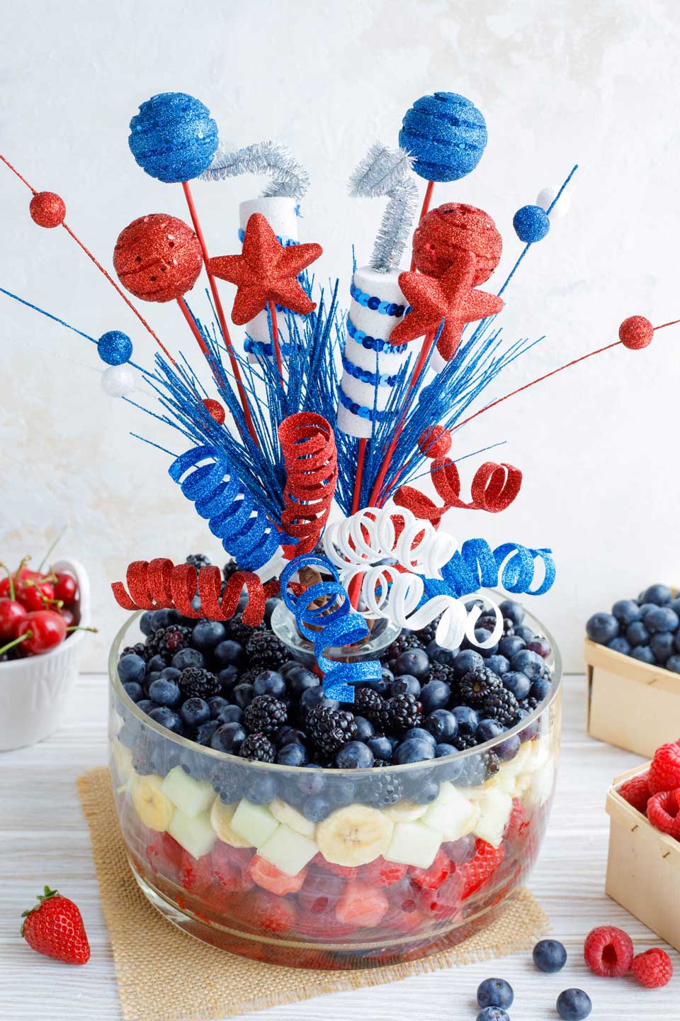 Centerpiece fruit salad featuring layered red white and blue fruits and festive party decorations coming up out of the middle of the bowl