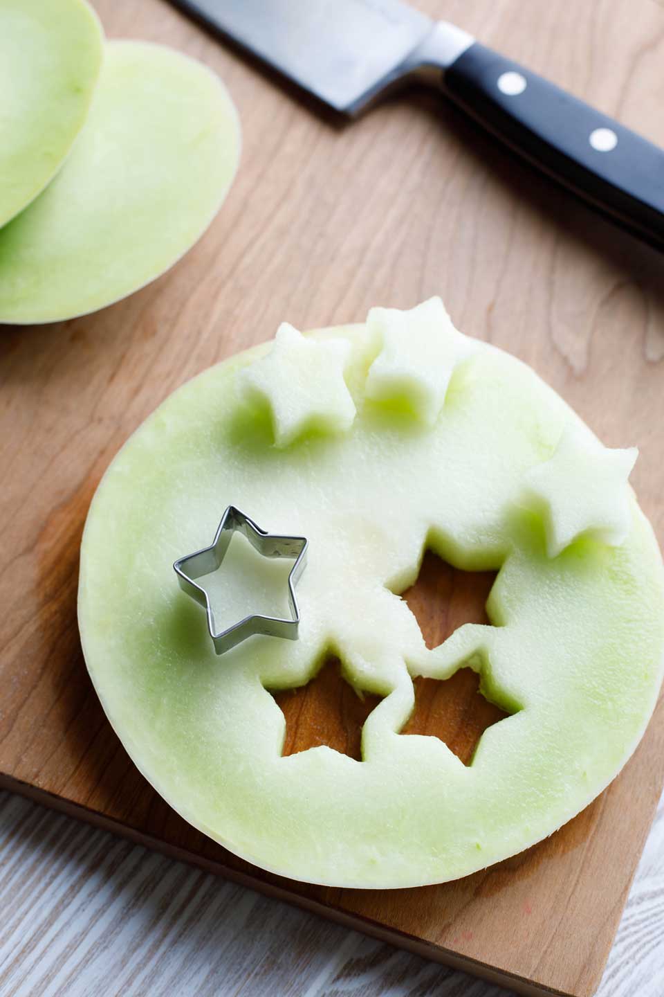 Slice of honeydew melon on a cutting board, with a small star-shaped cookie cutter making little melon stars to decorate the fruit salad