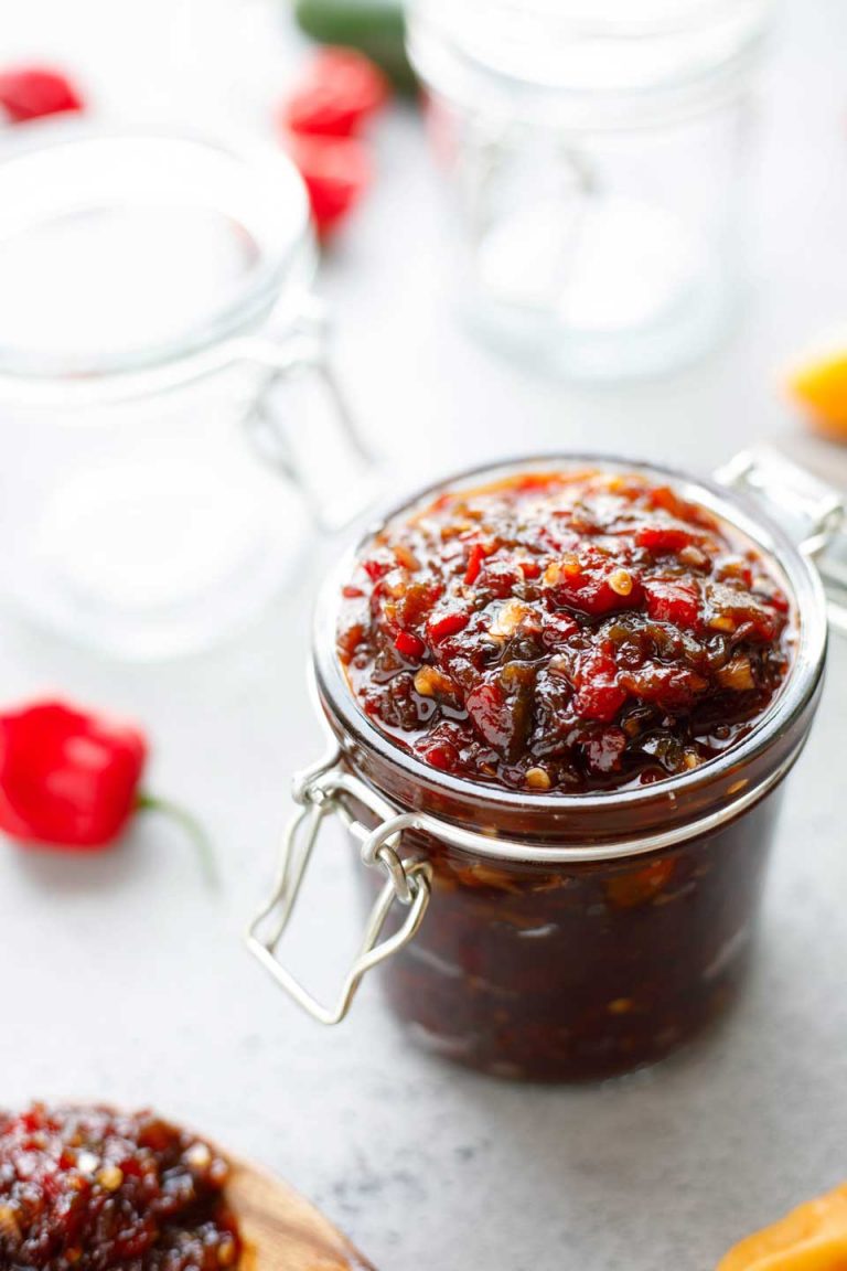 Glass jar full of relish with empty jars and raw, hot peppers in background.