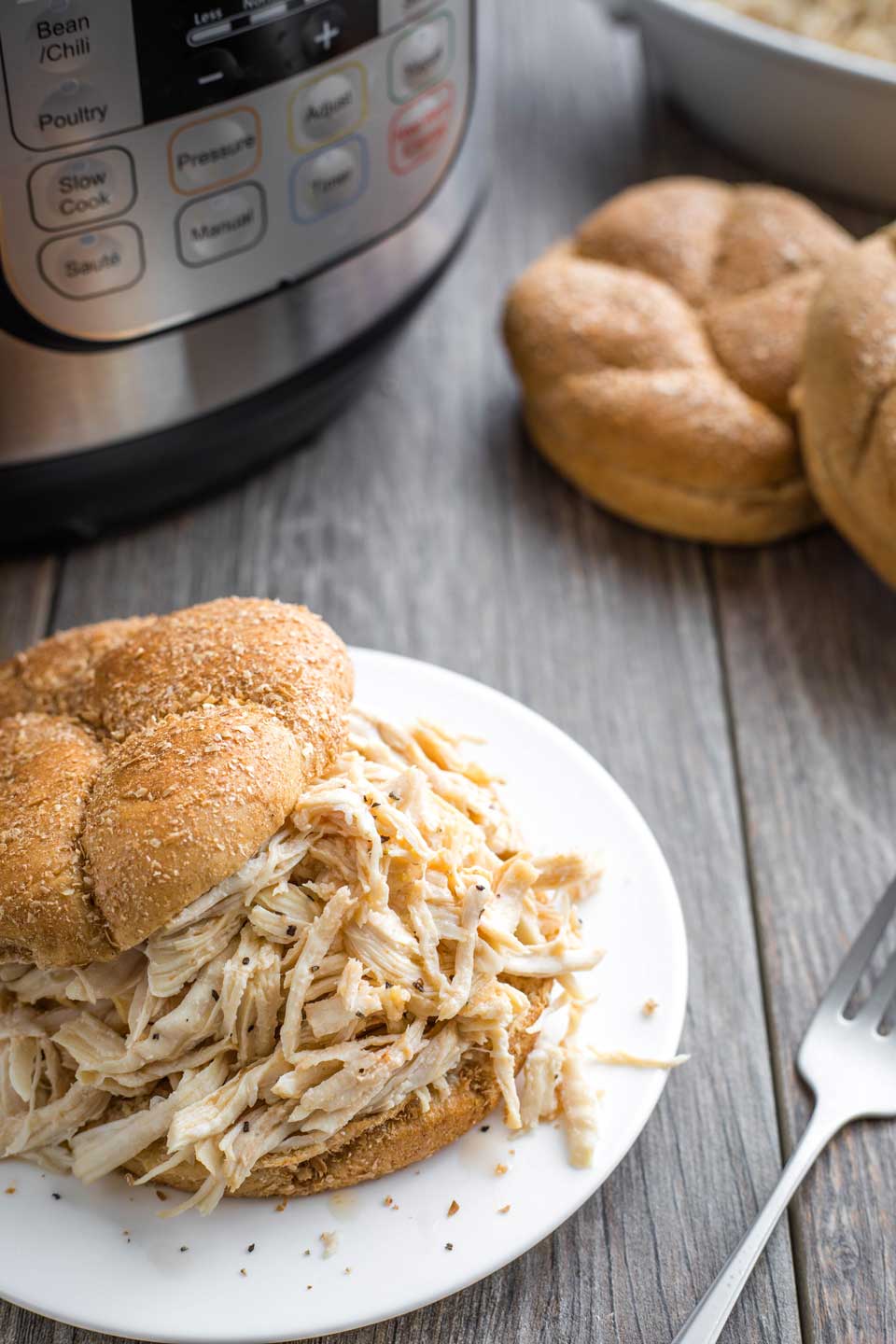 Instant Pot and buns in background with Instant Pot shredded chicken sandwich on a plate in the foreground