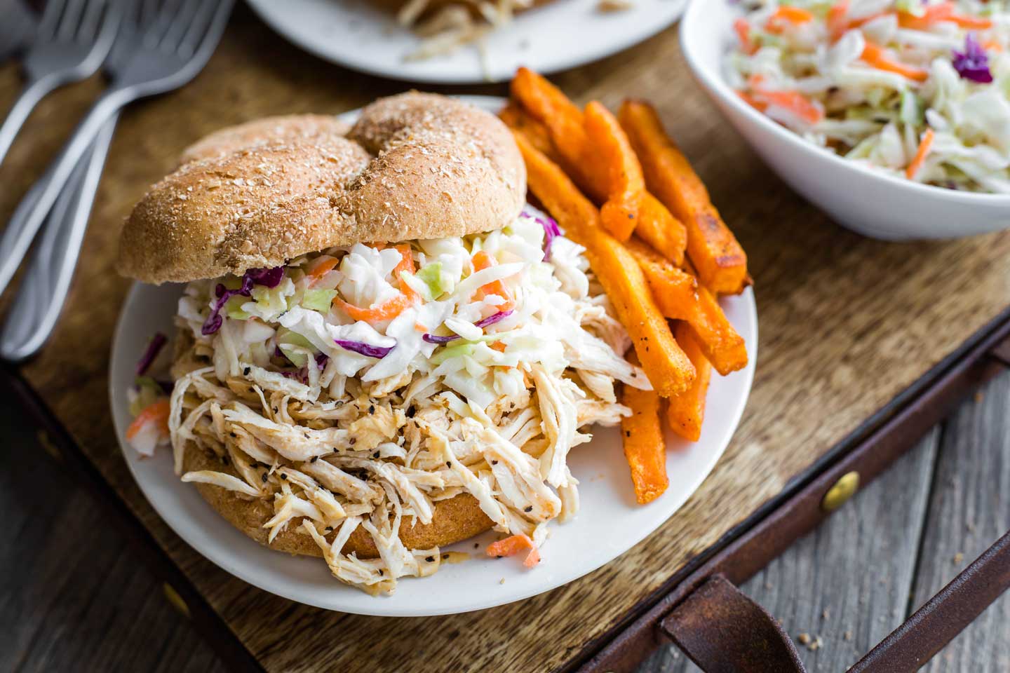 serving tray with extra forks and coleslaw, with a pulled chicken sandwich and sweet potato fries on a central plate