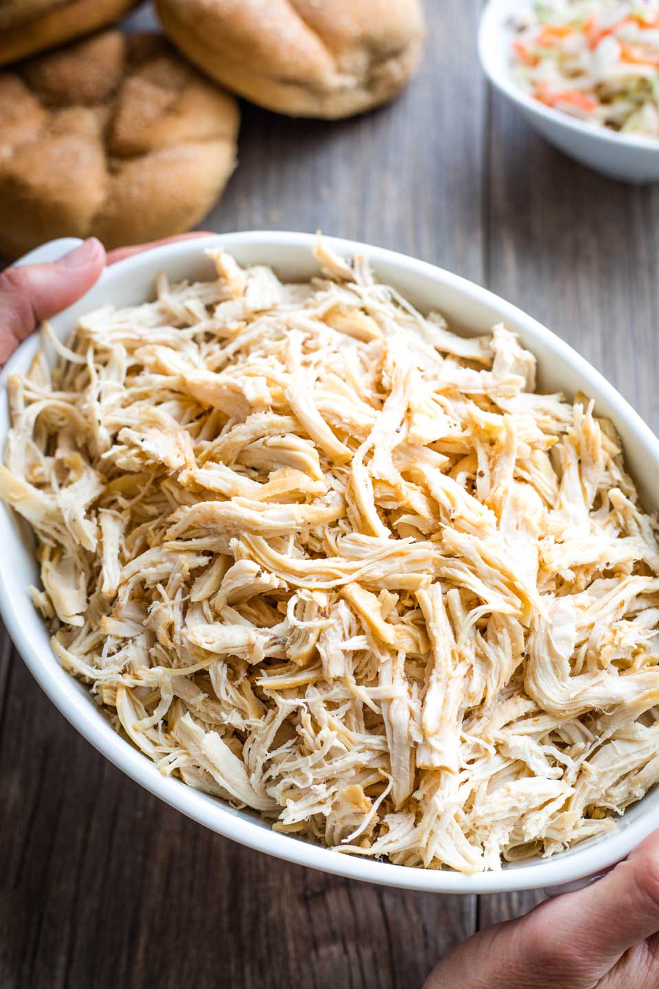 hands holding a serving bowl of shredded chicken, with buns and coleslaw in the background