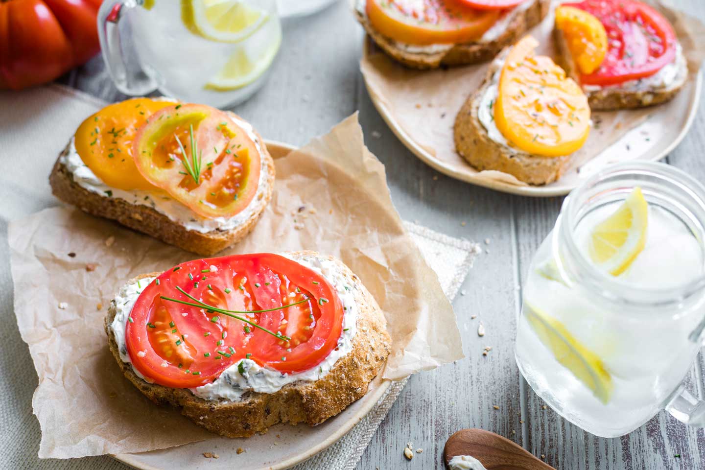 two open-faced tomato sandwiches on a plate, with others on platter in background and cold drinks at the side