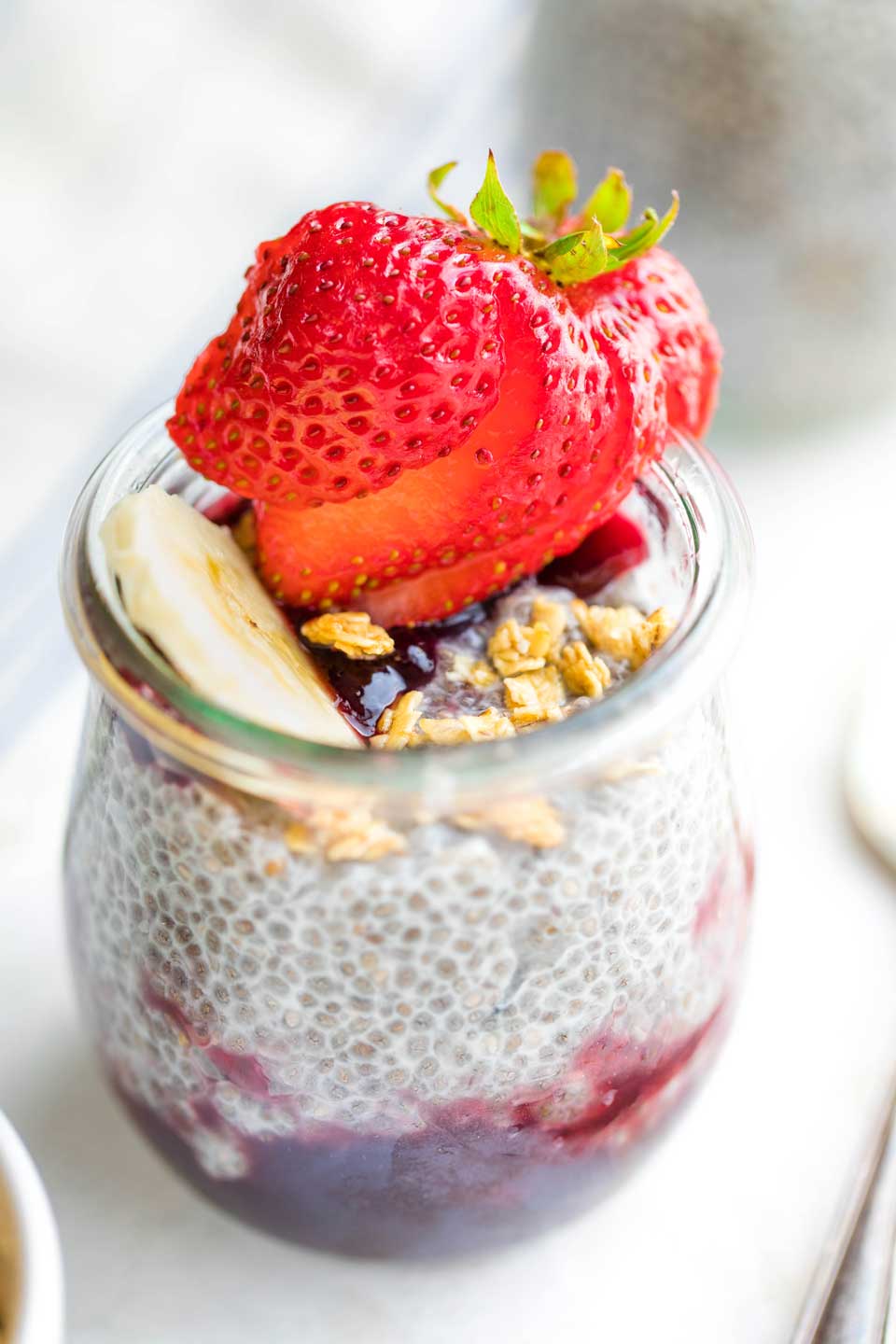 closeup of individual serving of chia pudding in glass "tulip" jar, topped with a strawberry and granola