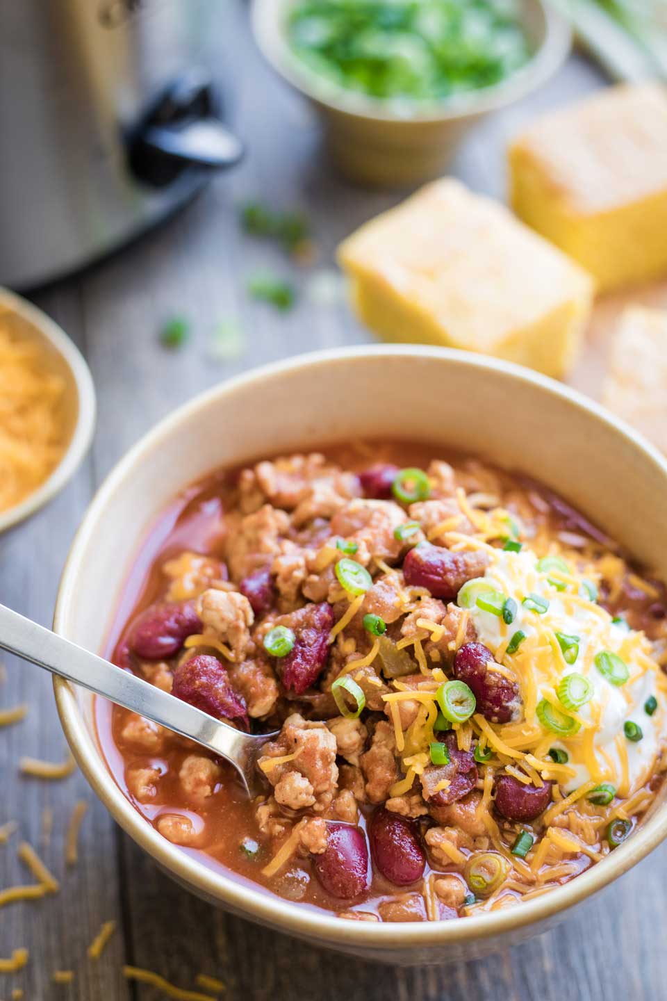 bowl of slow cooker chili, with toppings and a spoon - crock-pot and corn bread in background