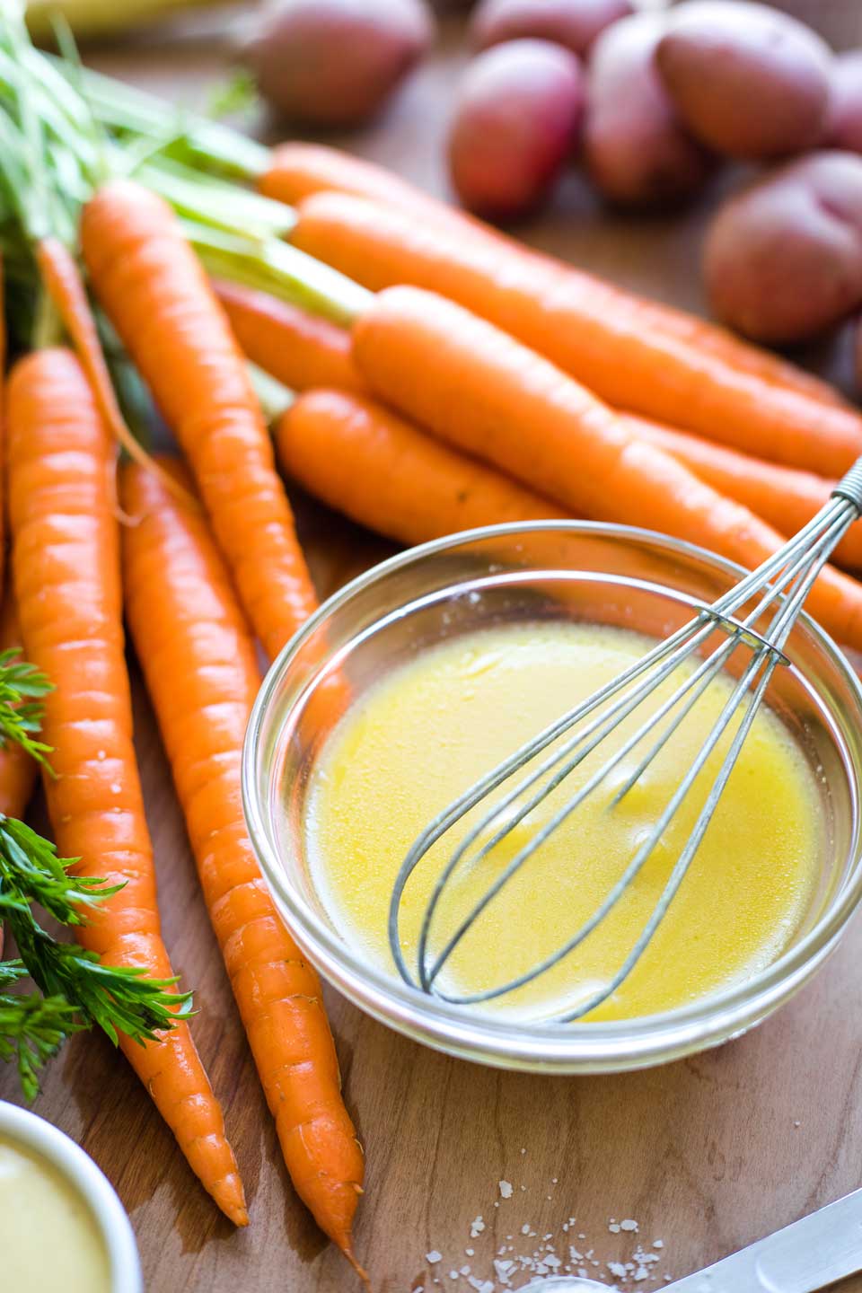 small bowl of Honey-Dijon Drizzle with whisk, nestled among raw carrots on cutting board