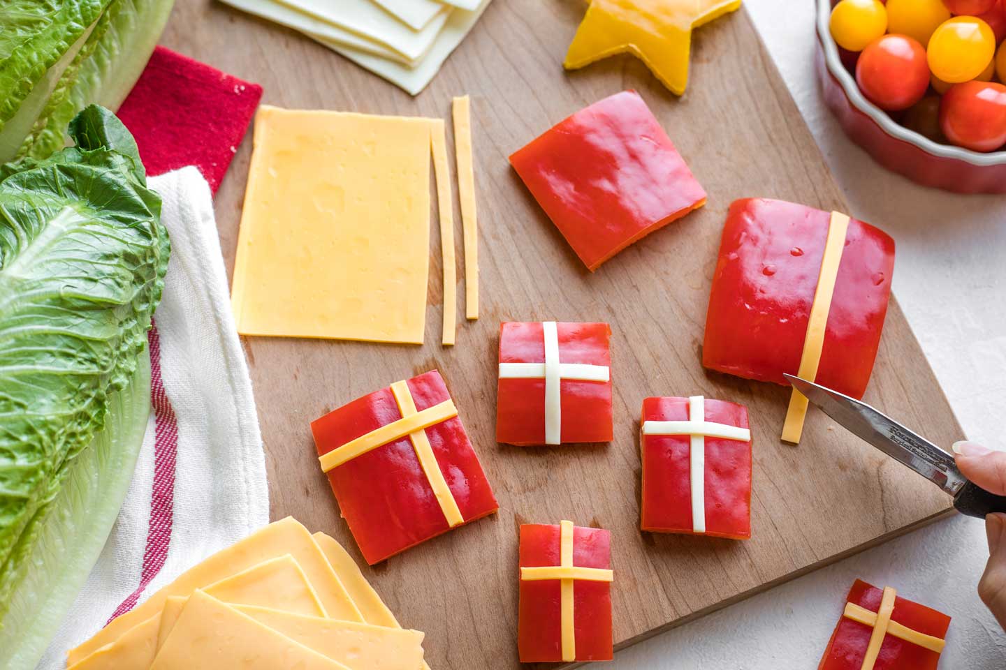 several square and rectangular pieces of red pepper on cutting board, being topped with thin strips of cheese to look like Christmas presents