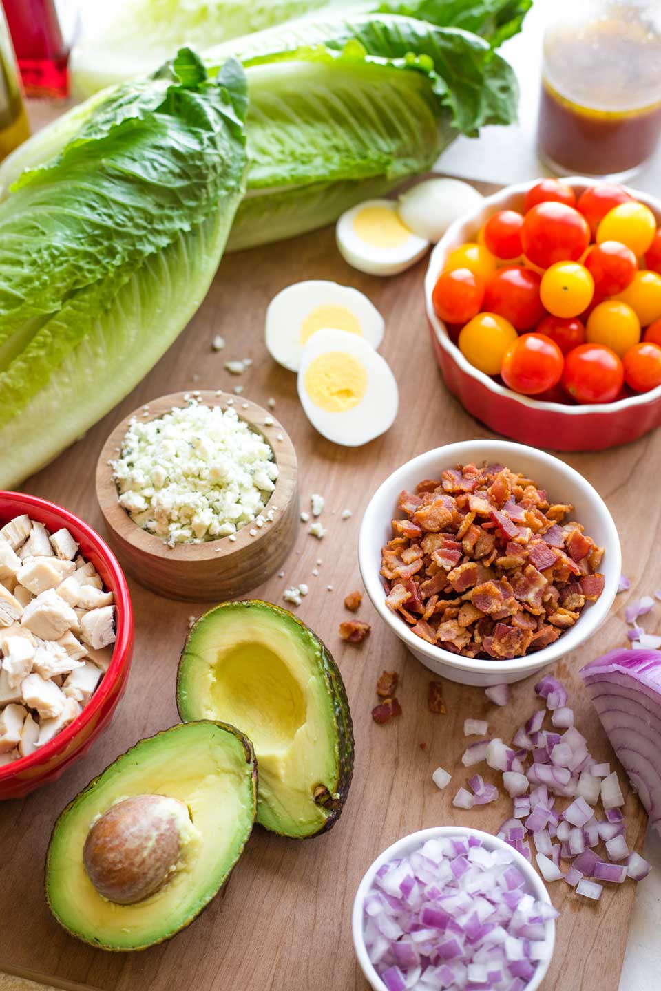 cutting board with prepped chicken Cobb Salad ingredients - romaine lettuce, hard-boiled eggs, cooked bacon, red onion, chicken, avocado and cherry tomatoes