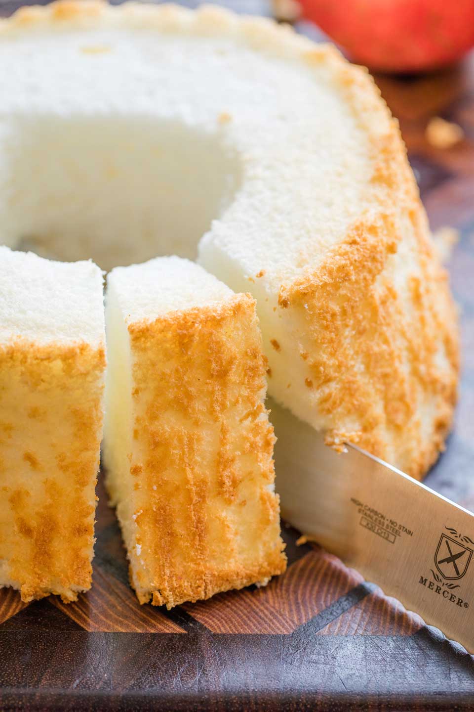 angel food cake on wooden cutting board, with one slice being cut with larger, serrated kinfe