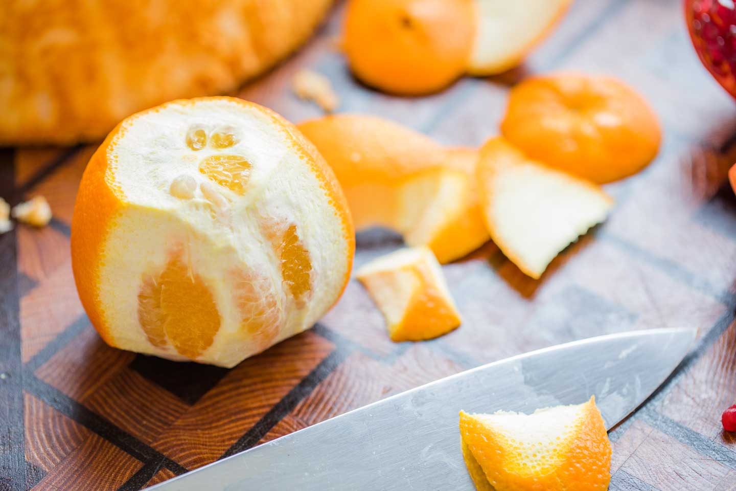 an orange on a wooden cutting board, with a chef's knife that has just started removing the orange peel through vertical cuts