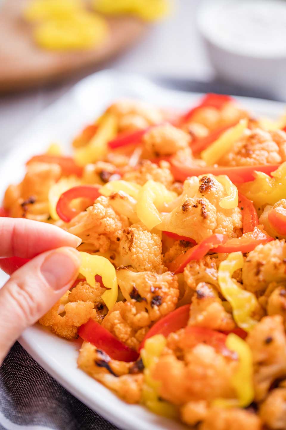 side view of white platter piled with buffalo cauliflower, mixed together with red peppers and banana peppers, with a hand at the edge adding one more banana pepper