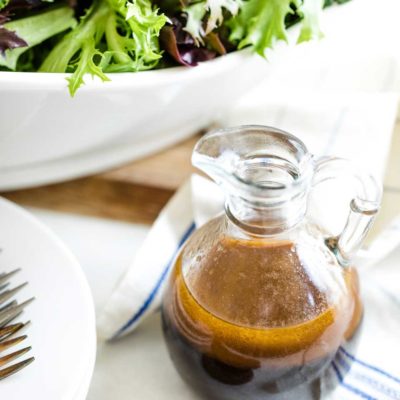 Cruet of dressing nestled near tea towel with navy stripe, with large bowl of salad, stack of plates and salad forks nearby