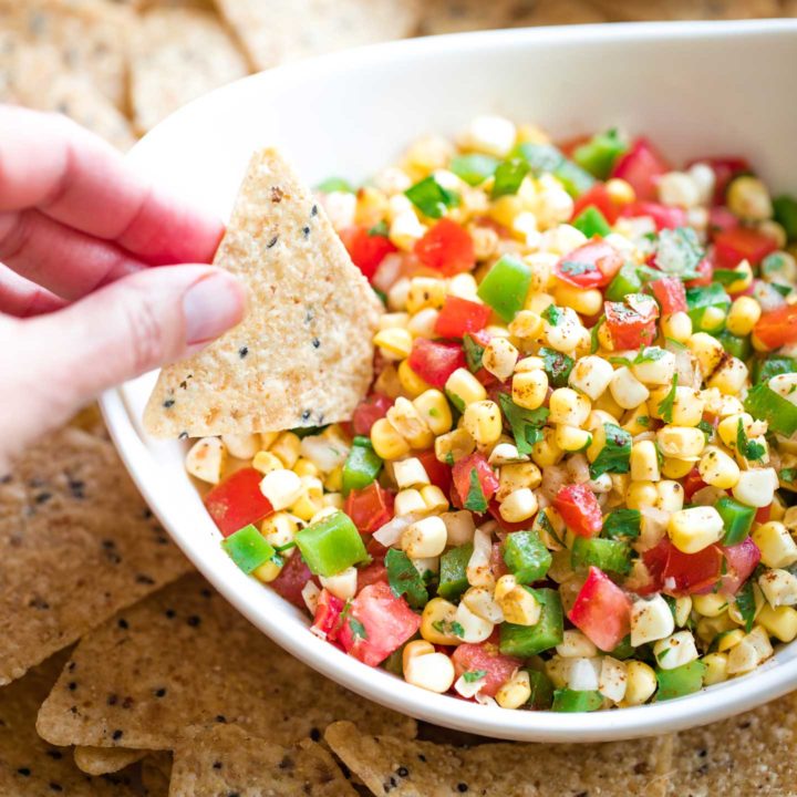 Large bowl of the corn salsa surrounded by tortilla chips, with a hand dipping one chip straight into the bowl