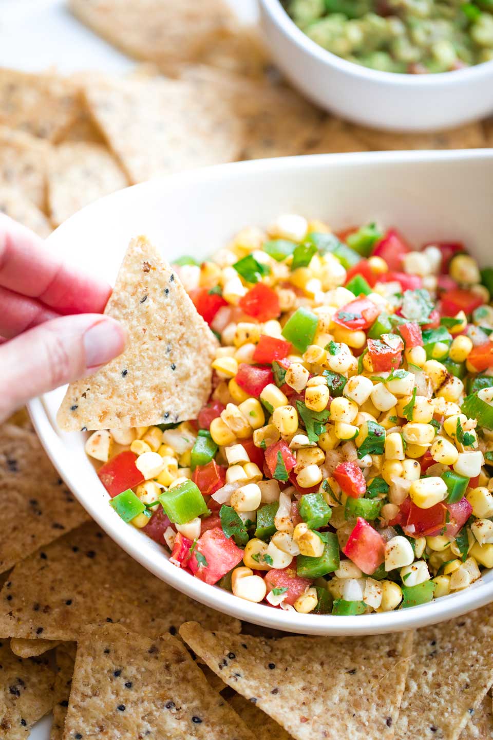 serving bowl filled with corn salsa, surrounded by whole grain tortilla chips, with a hand dipping one chip into salsa