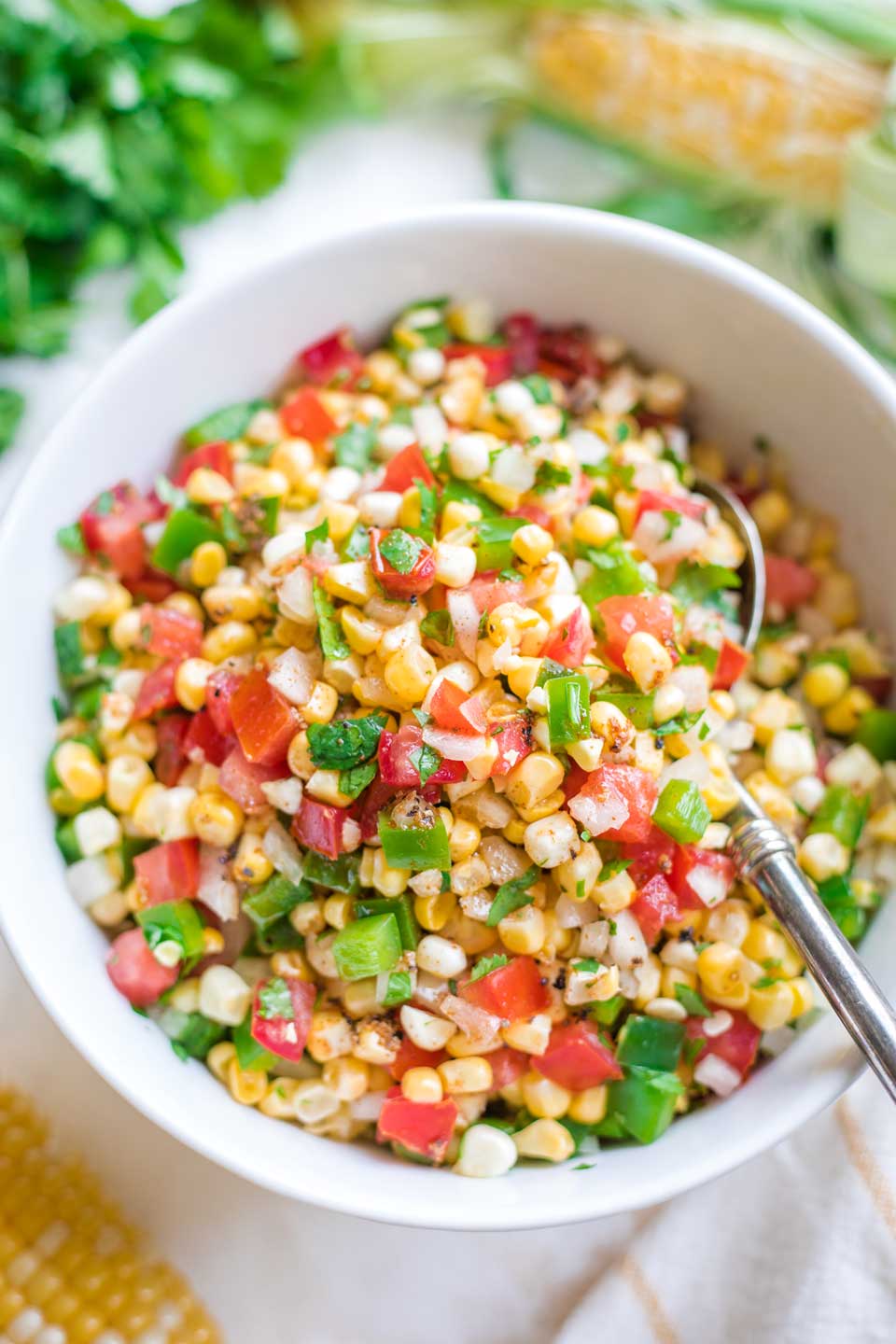 overhead photo of the salsa in a serving bowl with a spoon in it, with extra ears of corn and cilantro alongside