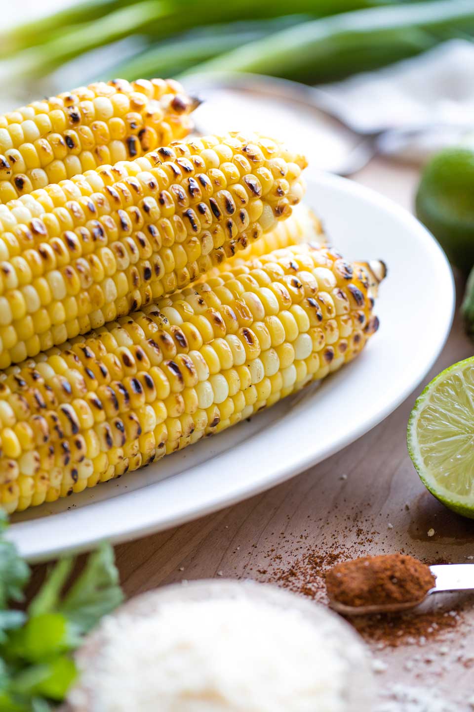A pile of 4 or 5 ears of grilled corn on a white platter, surrounded by other ingredients for this Mexican Corn Salad, like cut limes, cilantro, green onions and a spoonful of chile powder..