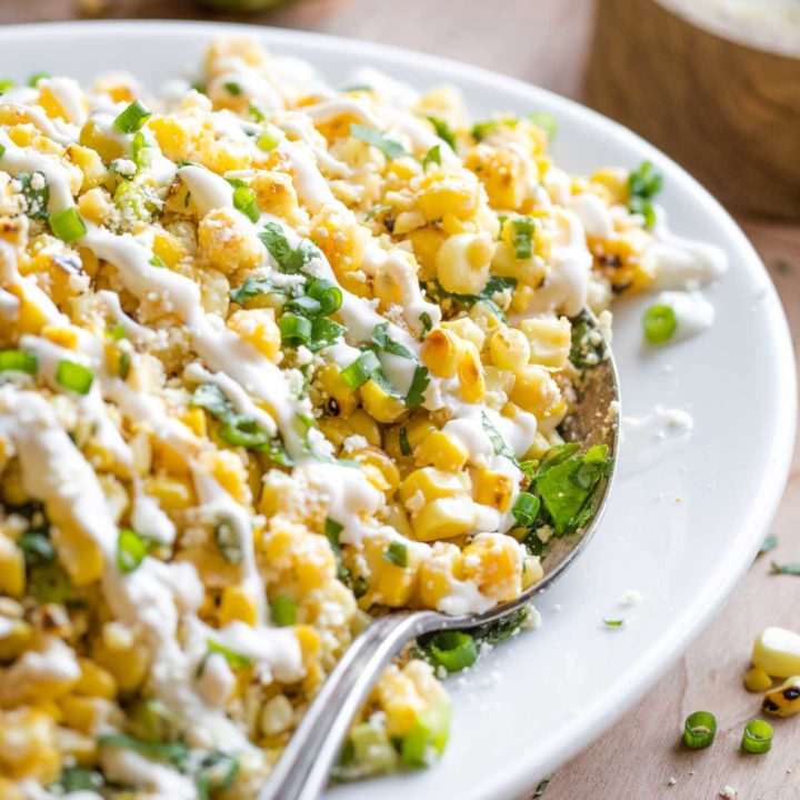 A closeup of a serving spoon tucked into the edge of this corn salad, which is presented on a white serving platter.