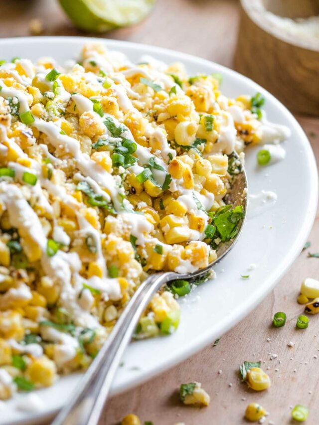 Closeup of about half the serving platter of Corn Salad, with a serving spoon dipped into the side, and squeezed lime rinds, extra cheese and cilantro leaves in the background.