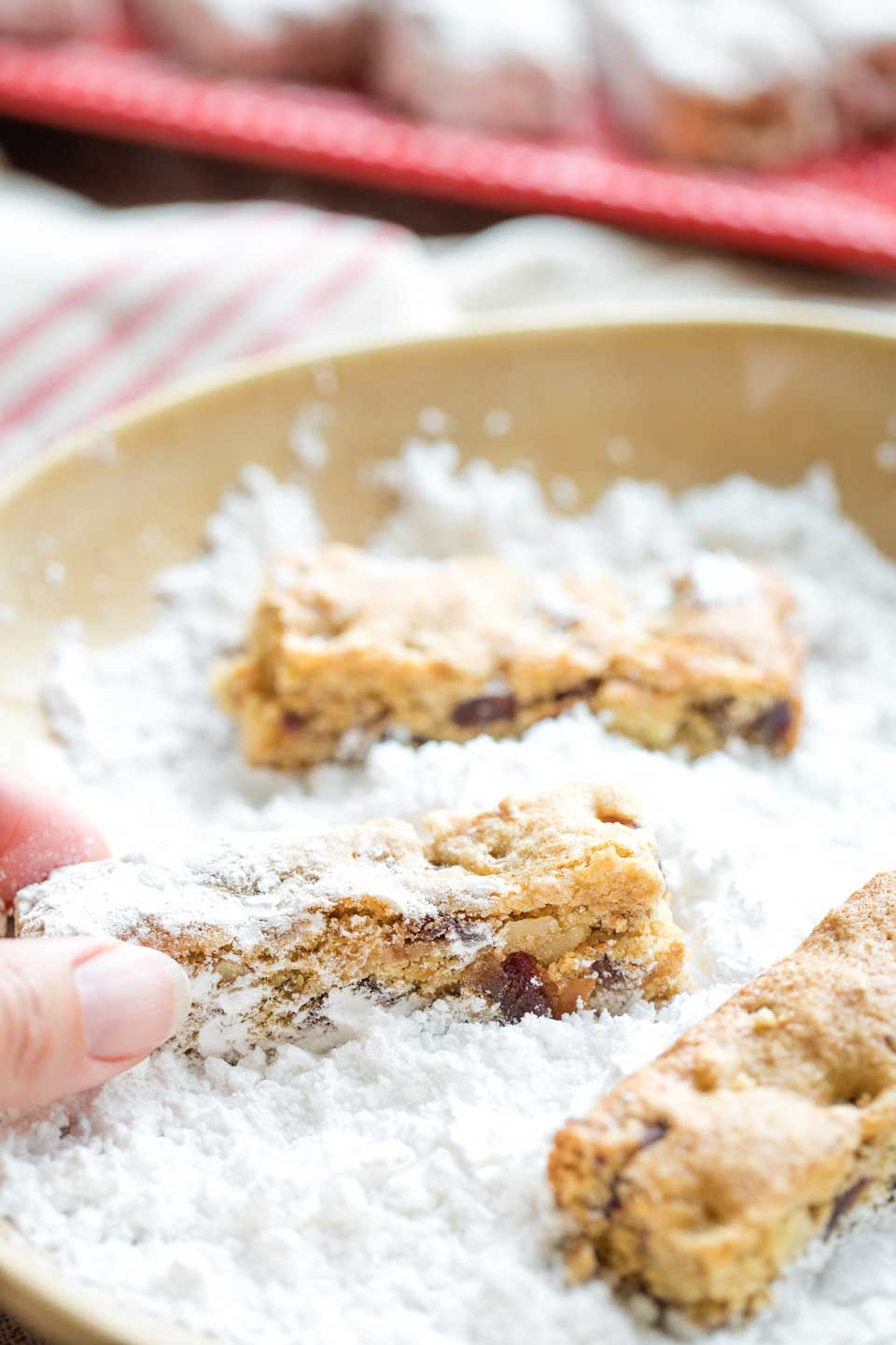 Closeup of three Date Bars in a bowl of powdered sugar, with fingers reaching in to roll one around in it.