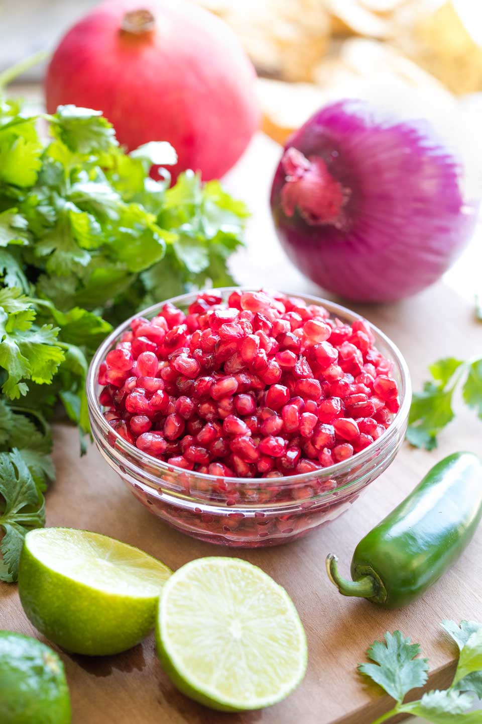 Ingredients for Pomegranate Salsa arranged on a cutting board, including a small bowl of arils, limes, a jalapeno, a bunch of cilantro, and a red onion.