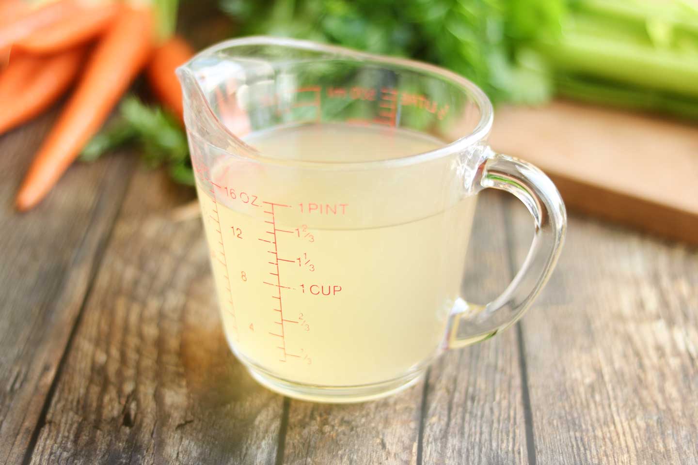 A 2-cup glass measuring cup full of chicken broth, with other ingredients in the background, ready to make this easy chicken noodle soup.