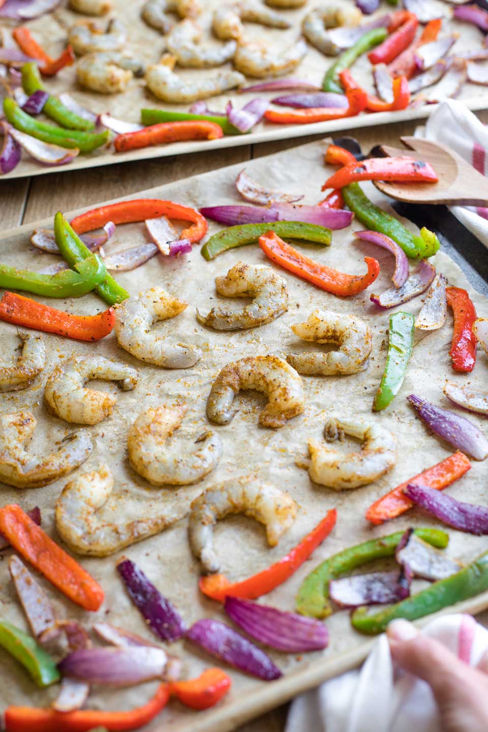 Seasoned raw shrimp being added to the sheet pans after vegetables have begun roasting.