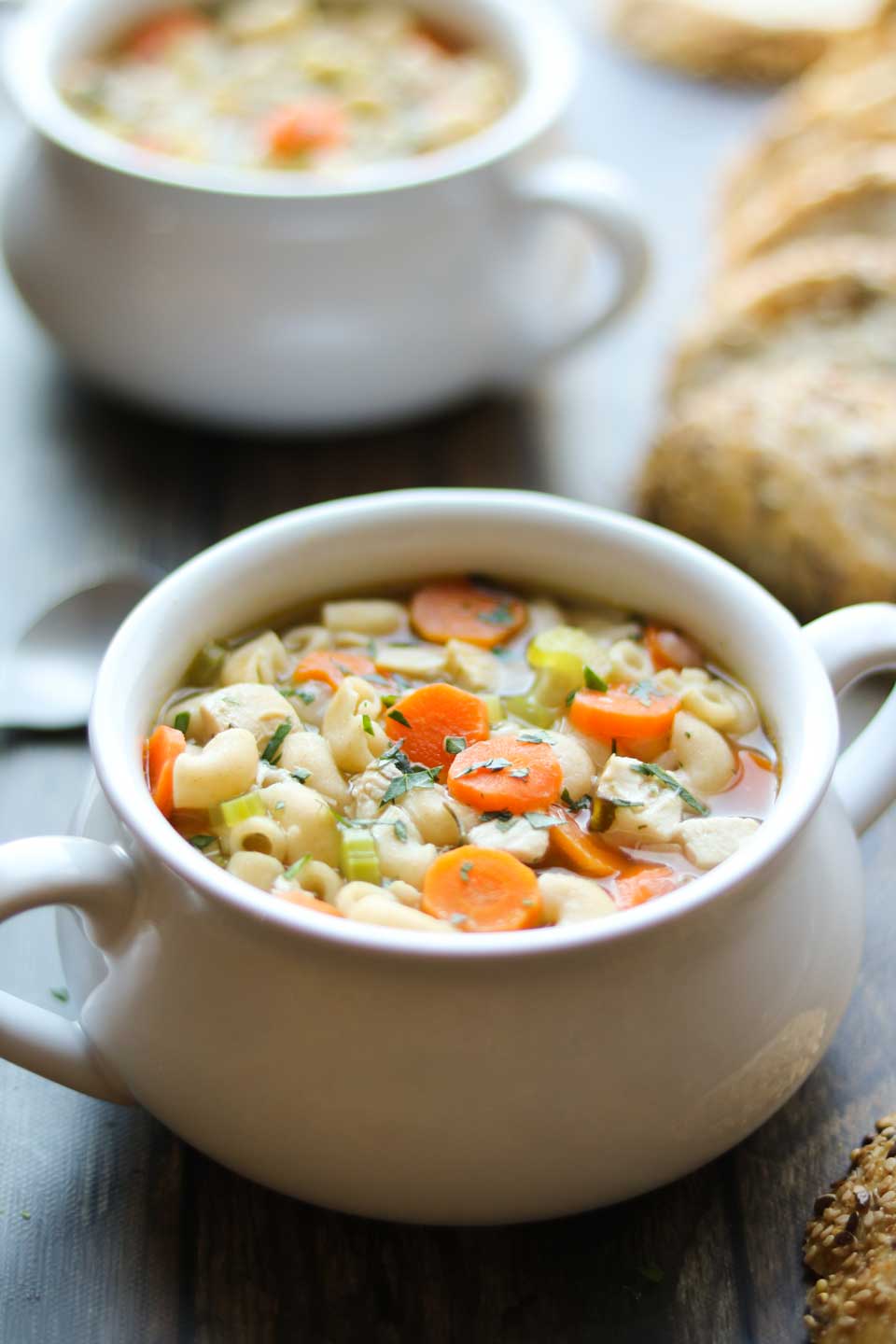 Hero photo of this roasted chicken noodle soup recipe, with a full bowl in the foreground and another in the background, with slices of granulated bread and a visible spoon on the sides.