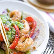 Closeup of one of these fajitas on a plate, so you can see the texture of the shrimp and peppers, with guacamole and sprinkled cheese on top and a fork in the foreground.