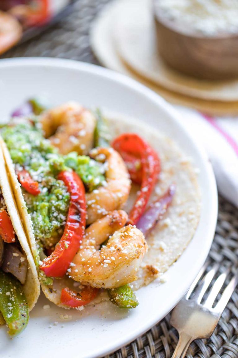 Closeup of one of these fajitas on a plate, so you can see the texture of the shrimp and peppers, with guacamole and sprinkled cheese on top and a fork in the foreground.