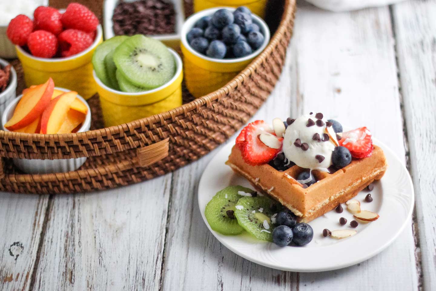 One loaded-up Galentine's Day waffle, piled high with various fruits, sprinkles and ice cream, sitting on a white plate with a basket filled with additional little bowls of toppings behind it.