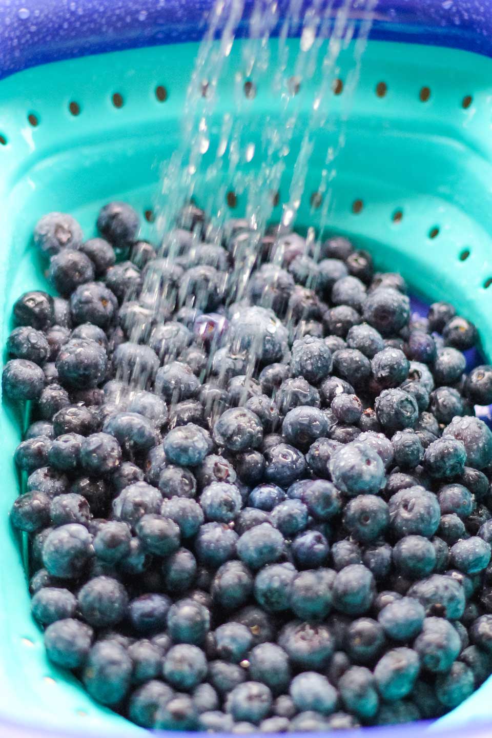 Big pile of fresh blueberries in a turquoise and blue strainer, being rinsed under a stream of water.