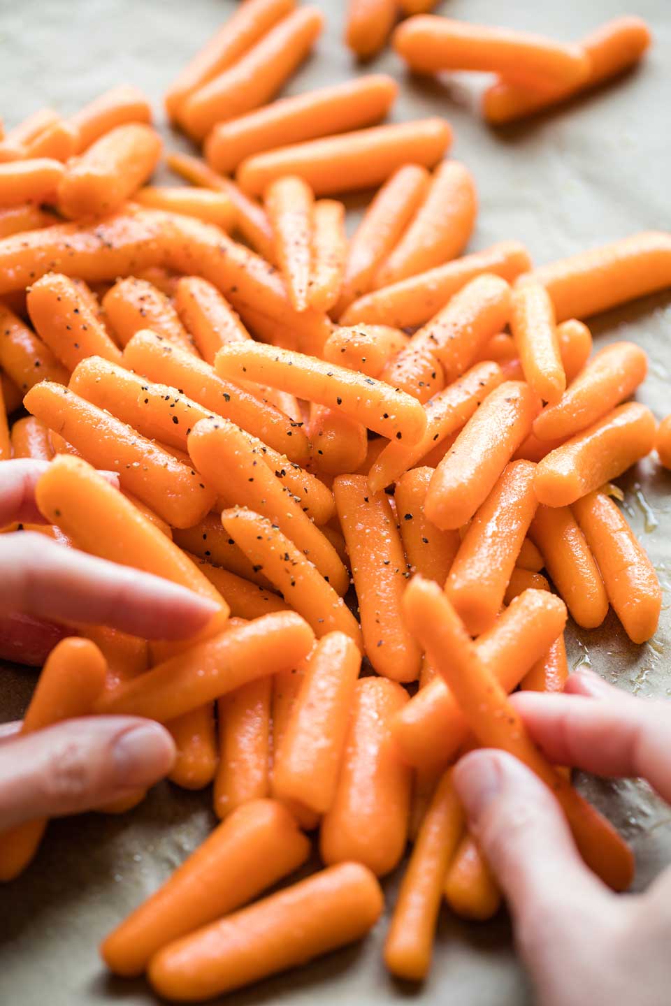 Two hands tossing raw baby carrots with oil and seasonings on a parchment-lined baking sheet before roasting in the oven.