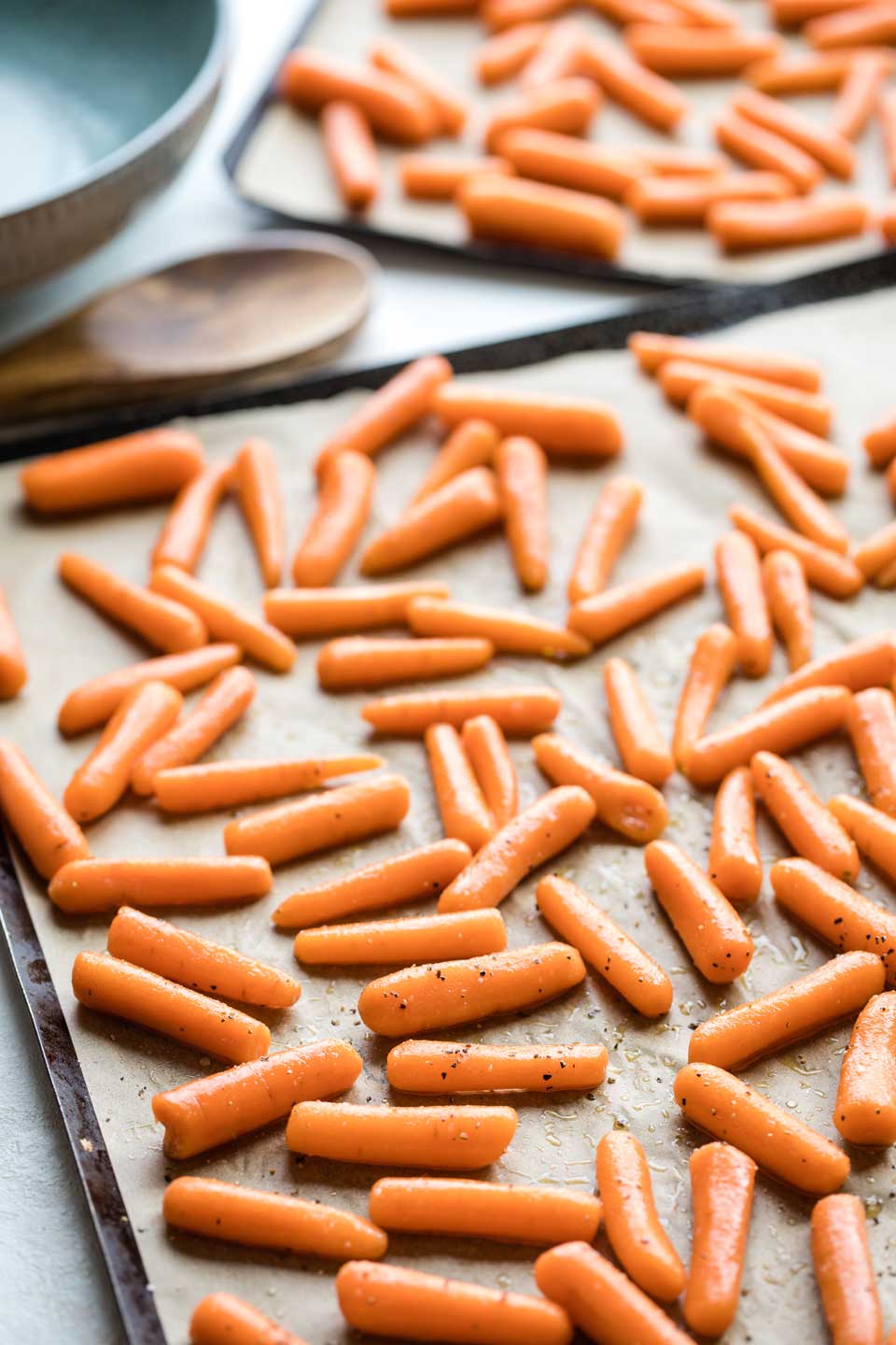 One parchment-lined roasting sheet (with another in the background), with raw, seasoned baby carrots spread out and ready to be put in the oven.