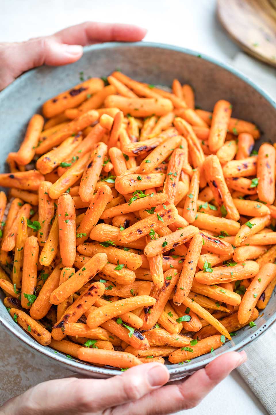 Overhead of two hands holding a blue serving bowl piled with roasted carrots that are fresh from the oven, ready to serve with a light sprinkling of fresh parsley.