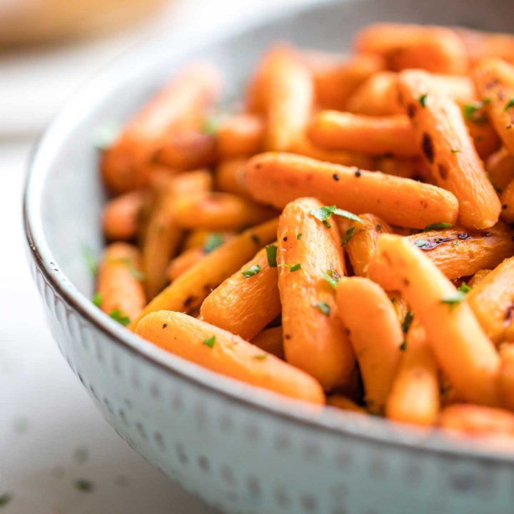 Closeup of one side of a blue serving bowl, piled with roasted carrots flecked with a sprinkling of chopped parsley.
