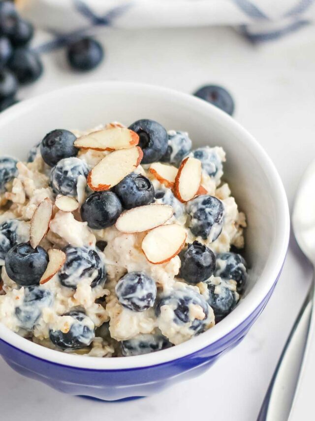 The finished recipe presented in a little blue bowl with a sprinkling of extra blueberries and sliced almonds on top of the cold oats mixture, with a spoon alongside..