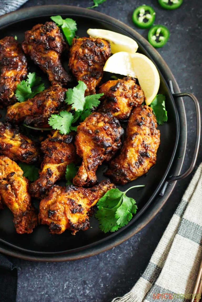 Partial overhead photo of a handled black serving tray of these wings, garnished with cilantro leaves and lemon wedges, with a black-striped towel alongside.