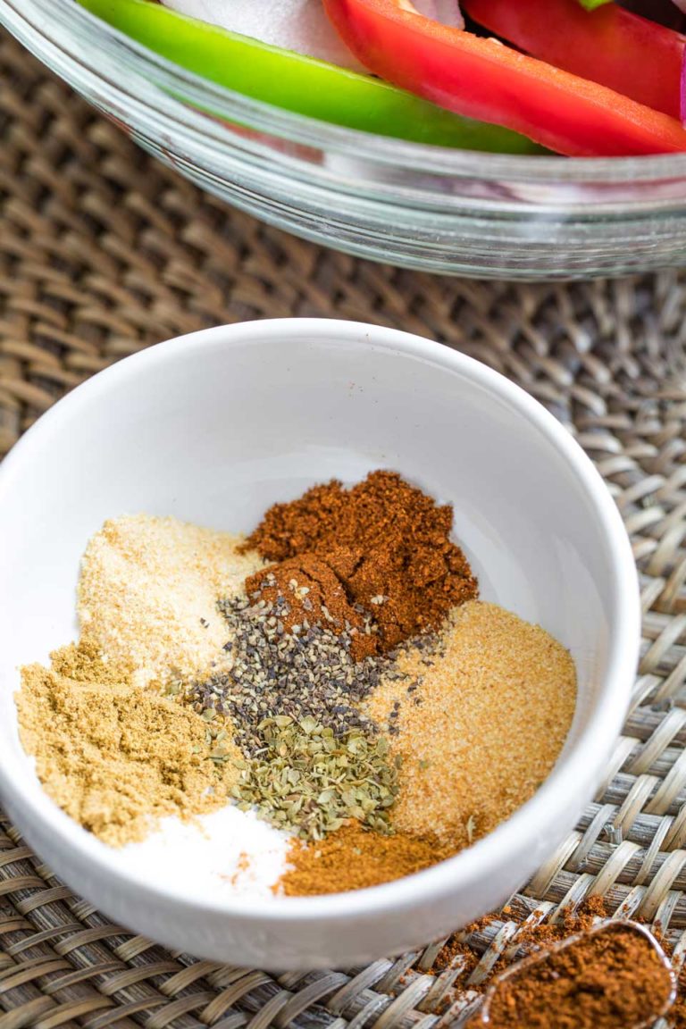 White mixing bowl containing spices before they're mixed together, with a teaspoonfull of spice nearby and a bowl of sliced fajitas veggies in the background.