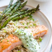 Closeup of the finished recipe, with the pan seared salmon arranged on a rimmed dinner plate, served with rice and asparagus, with utensils and additinal sauce just visible at the edges of the photo.