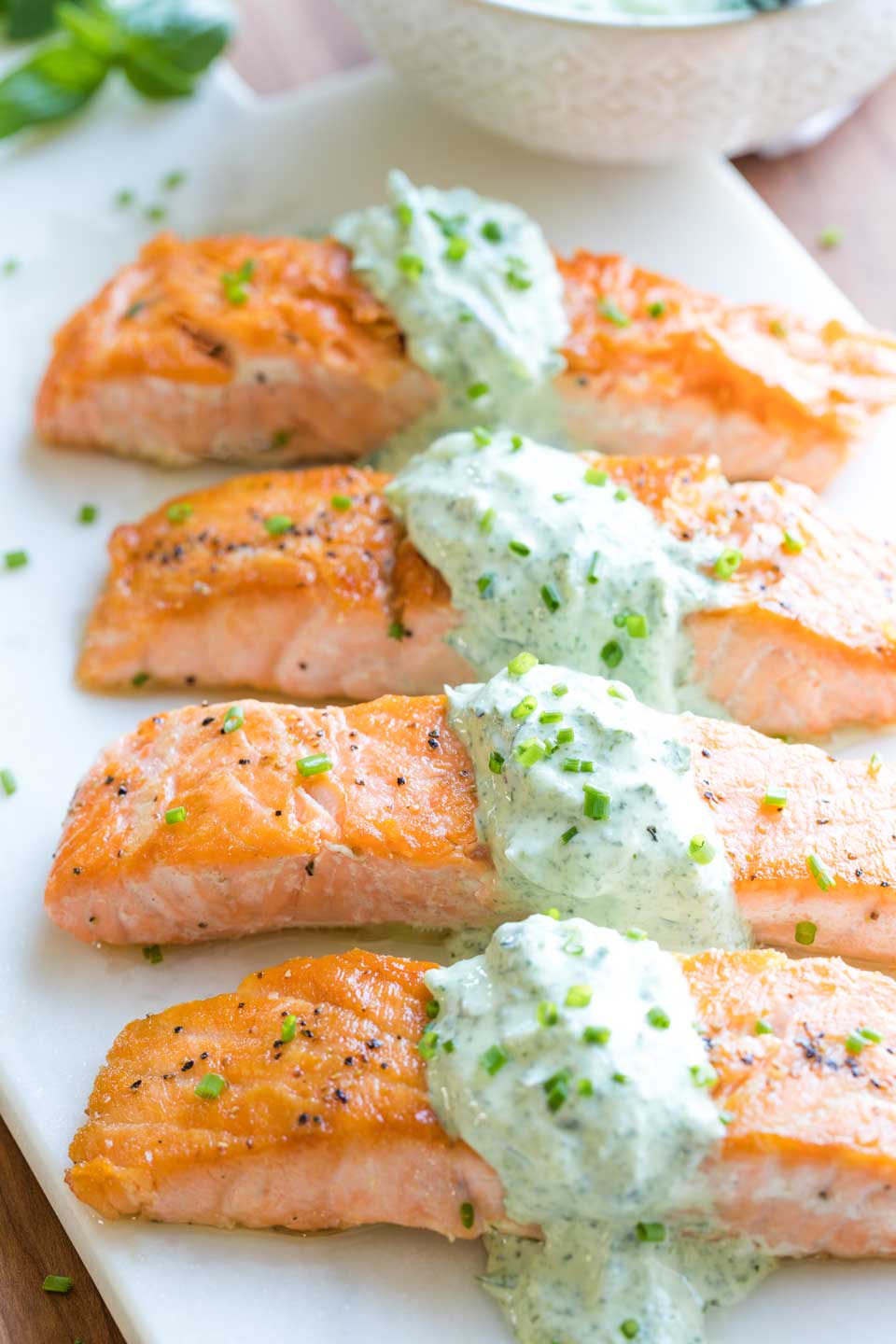Four salmon fillets served on a white tray, so you can see the pan seared top crust, draped across the center of each fillet with the tzatziki sauce and sprinkled with chopped chives for serving.