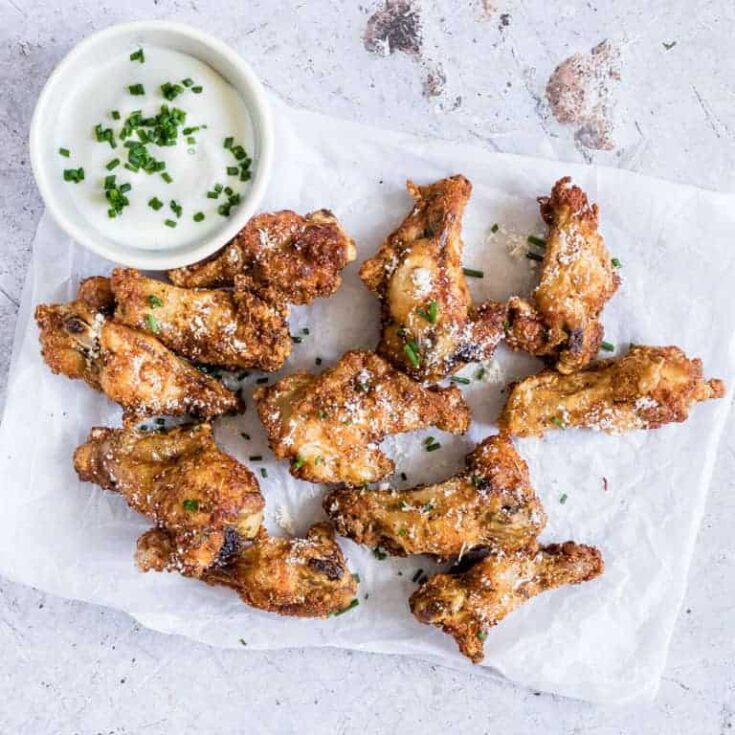 Batch of 11 air fryer wings laying on shite paper on a white background, with a small bowl of scallion-topped dip alongside.