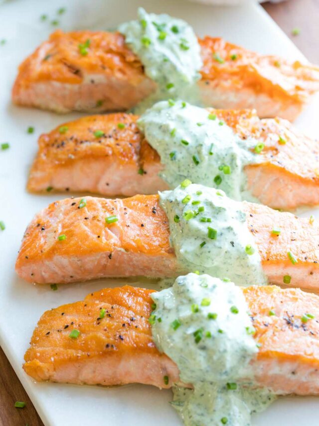 Four salmon fillets served on a white tray, so you can see the pan seared top crust, draped across the center of each fillet with the tzatziki sauce and sprinkled with chopped chives for serving.