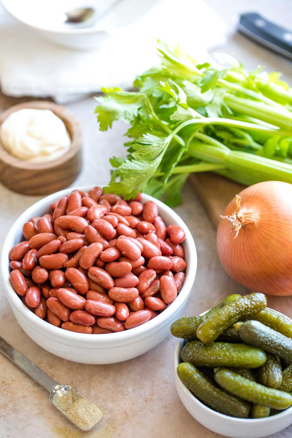 Ingredients for the salad: kidney beans and sweet pickles each in white bowls, an onion, stalks of celery, and a wooden bowl of mayo.